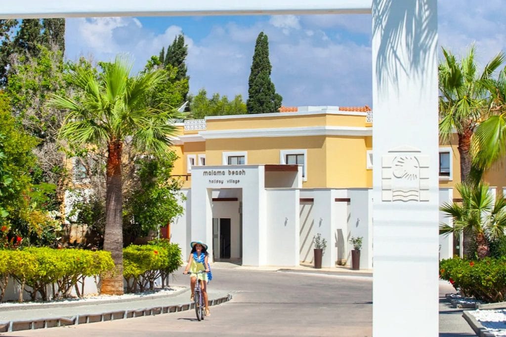 White entrance gateway with signage at Malama Beach Holiday Village, a guest cycling through, Protaras